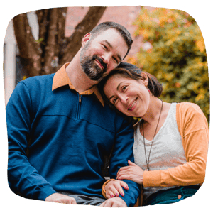 Middle aged couple smiling at the camera while sitting on a green bench outside-1
